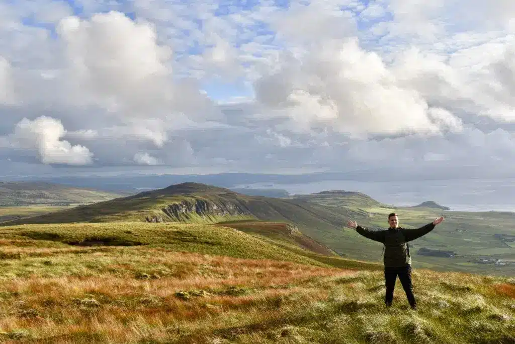 En vandrare med utsträckta armar på en gräsbevuxen bergsrygg på Skye